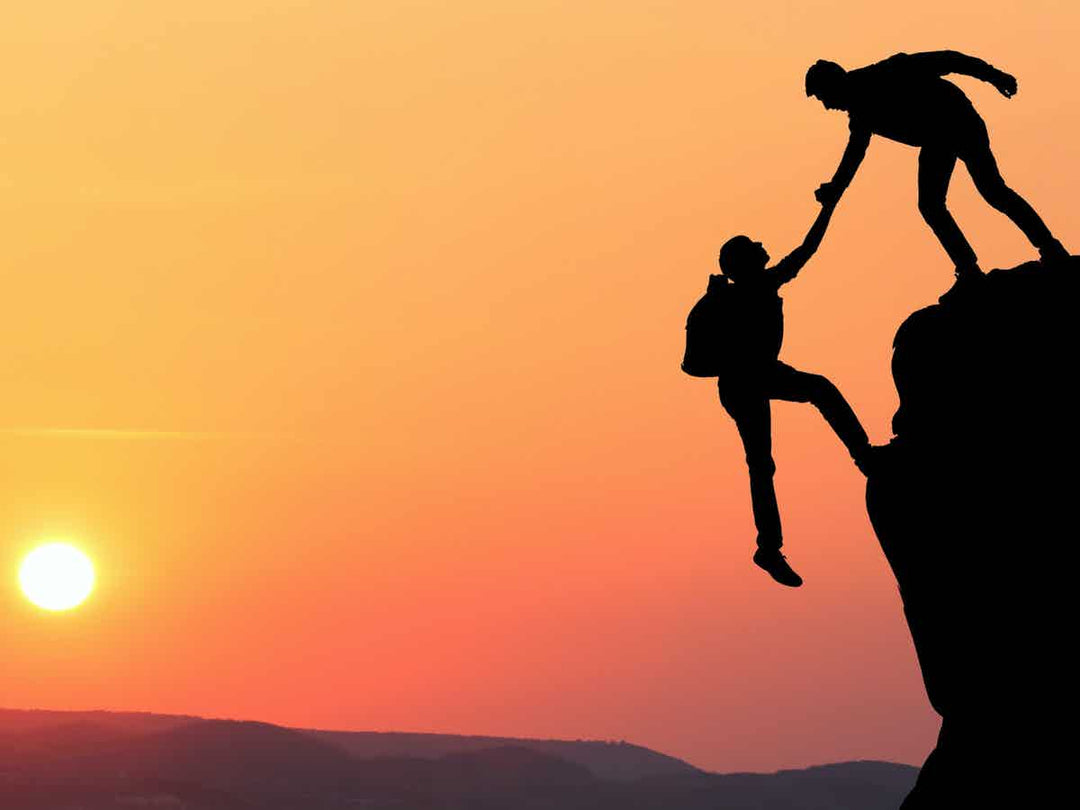 Person helping person up from cliff edge with setting sun in the background